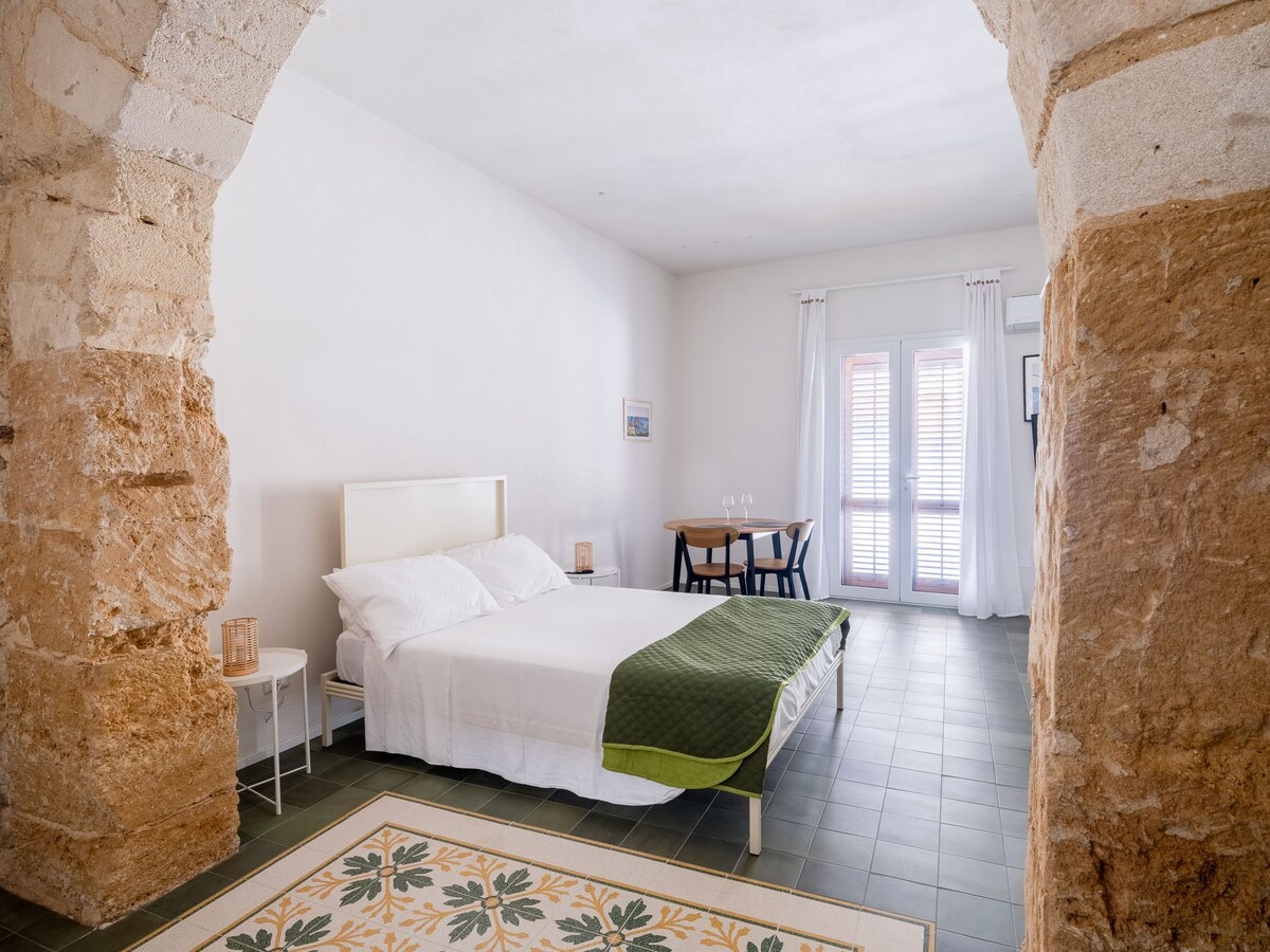 A spacious sleeping area features a large bed with a crisp white duvet and a green throw. Natural light fills the room through large windows, which are framed by white curtains. A dining table with chairs is positioned nearby, creating a cozy atmosphere.
