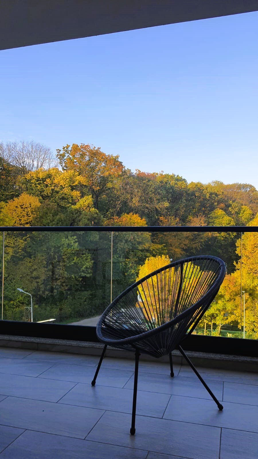 A private balcony showcases a black woven chair facing a panoramic view of the Băneasa Forest, adorned with autumn foliage. The clear blue sky complements the vibrant greens and yellows of the trees, creating a serene outdoor space for relaxation.