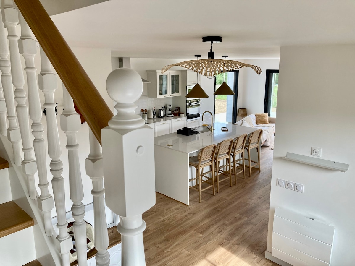 An open kitchen area is presented, featuring a central island with modern bar stools. Bright pendant lights hang above, illuminating sleek cabinetry and appliances. Large windows allow natural light to fill the space, enhancing the airy feel of the living area nearby.
