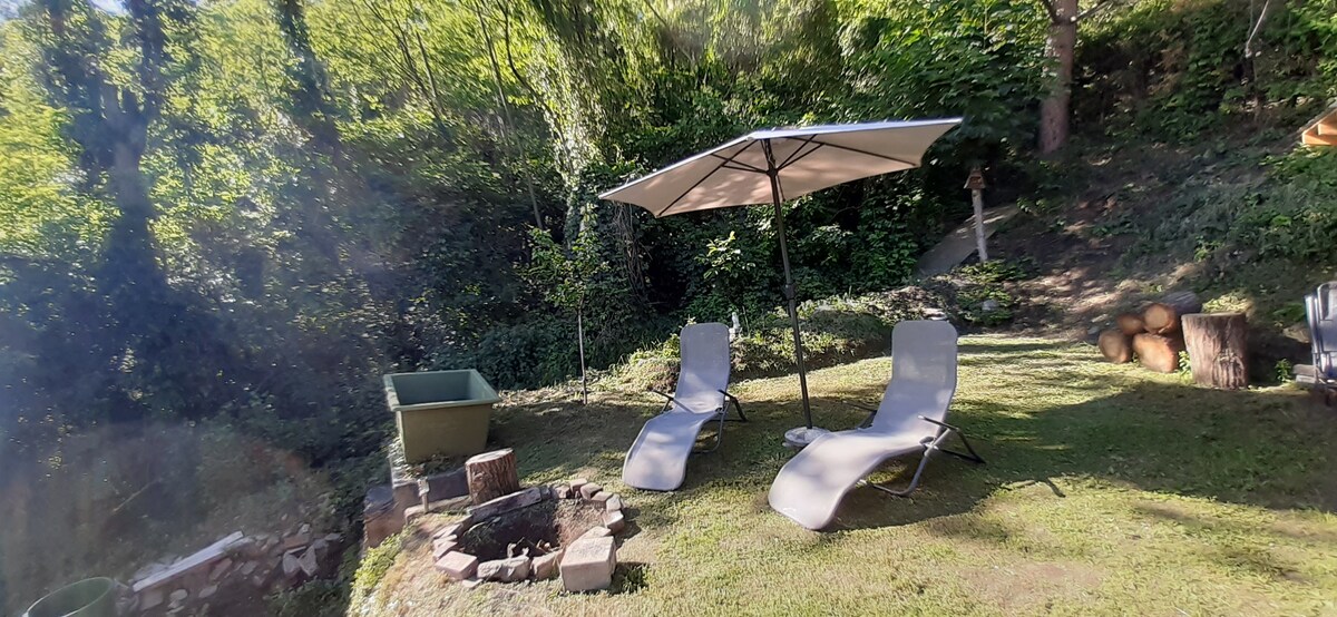 A peaceful garden area features two reclining lounge chairs under a large umbrella, providing shade. A fire pit, surrounded by a circular stone edge, is located nearby. Lush greenery and a small storage container are visible in the background, creating an inviting outdoor retreat.
