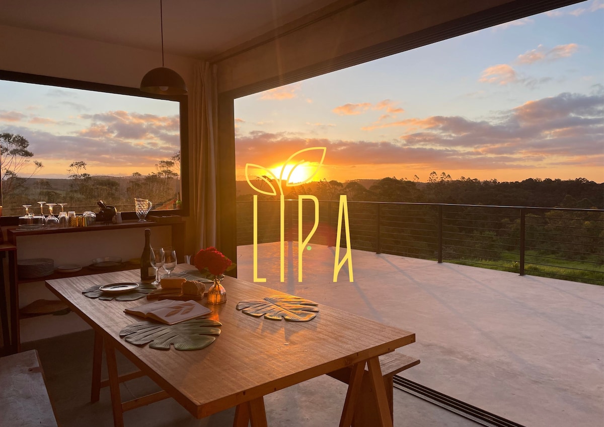 A dining table is set with dinnerware and flowers, positioned near large glass doors that frame a sunset view over lush greenery. The warm glow of the sun highlights the serene outdoor landscape while providing a harmonious indoor-outdoor atmosphere.