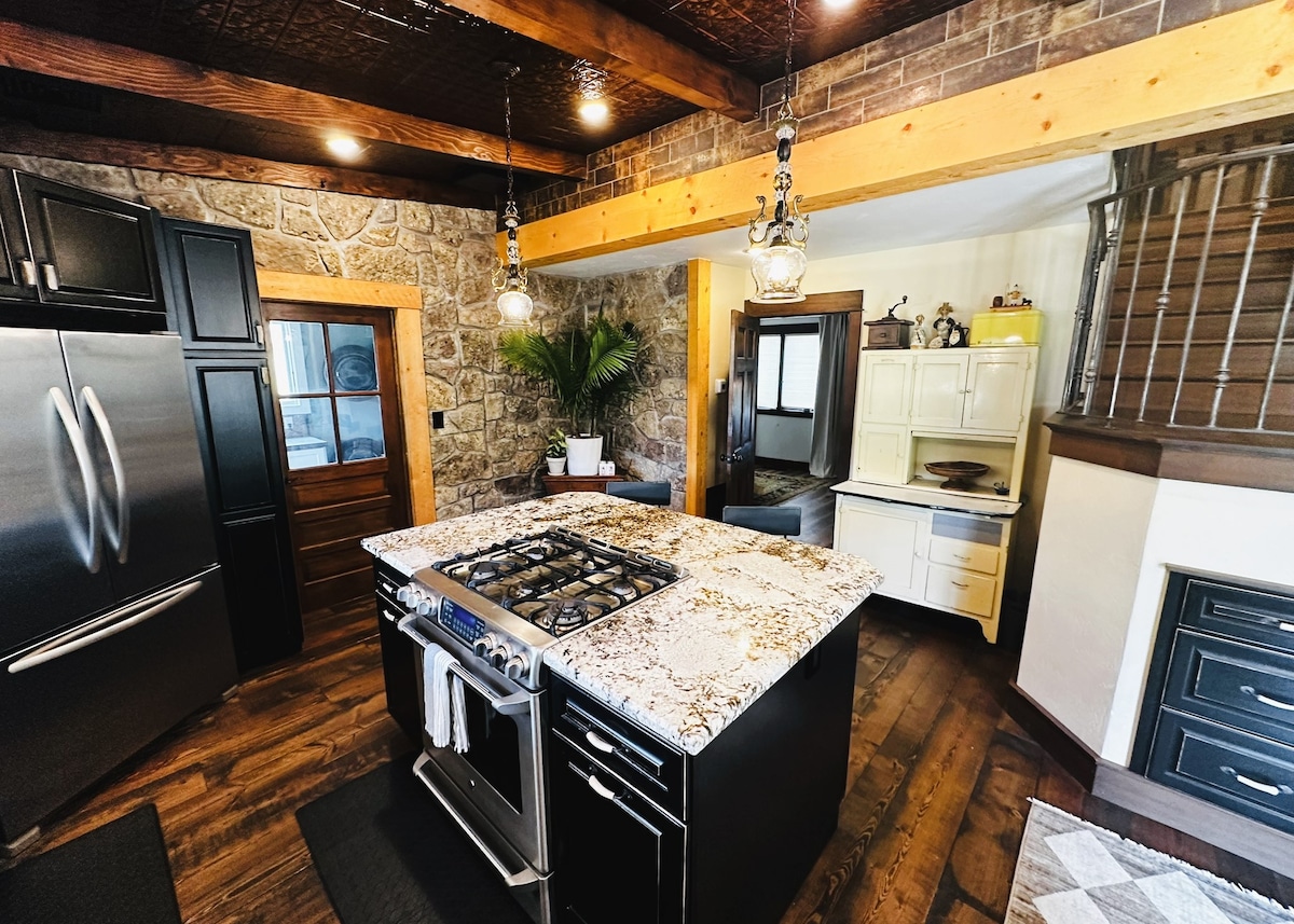 The kitchen features a large island with a stone countertop and a gas stove. Dark cabinetry contrasts with the rustic stone walls and wooden beams. Stainless steel appliances are present, and a potted plant adds a touch of greenery to the space.