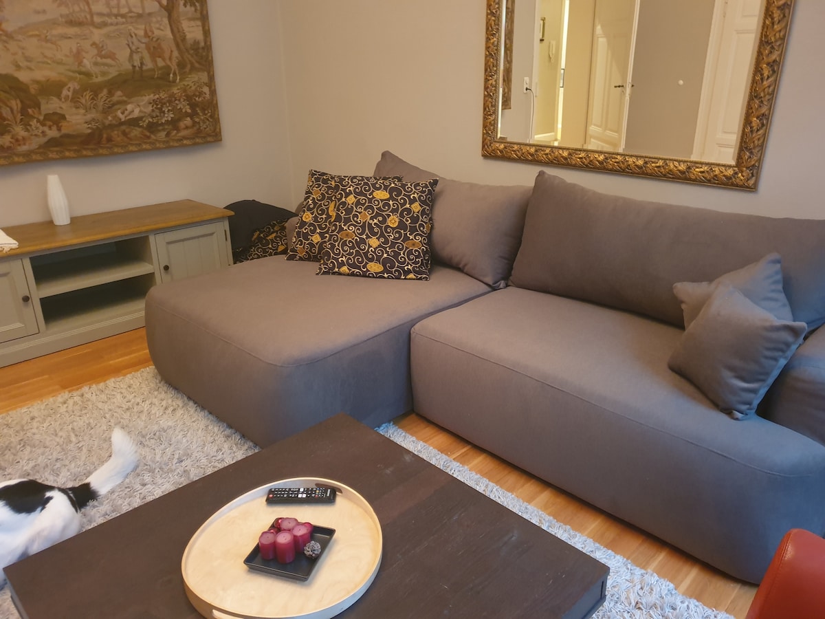 A spacious living area is shown, featuring a large, gray sectional sofa adorned with decorative cushions. A wooden coffee table holds a platter of candles, while a textured area rug adds warmth. The backdrop features a framed artwork and a light wooden cabinet.