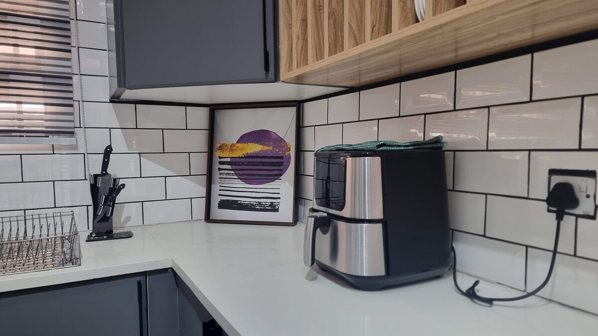 A modern kitchen corner displays a sleek air fryer alongside a framed piece of artwork featuring abstract colors. The white tiled backsplash contrasts with dark cabinetry, and utensils are organized neatly in a stand on the countertop, contributing to the functional design.