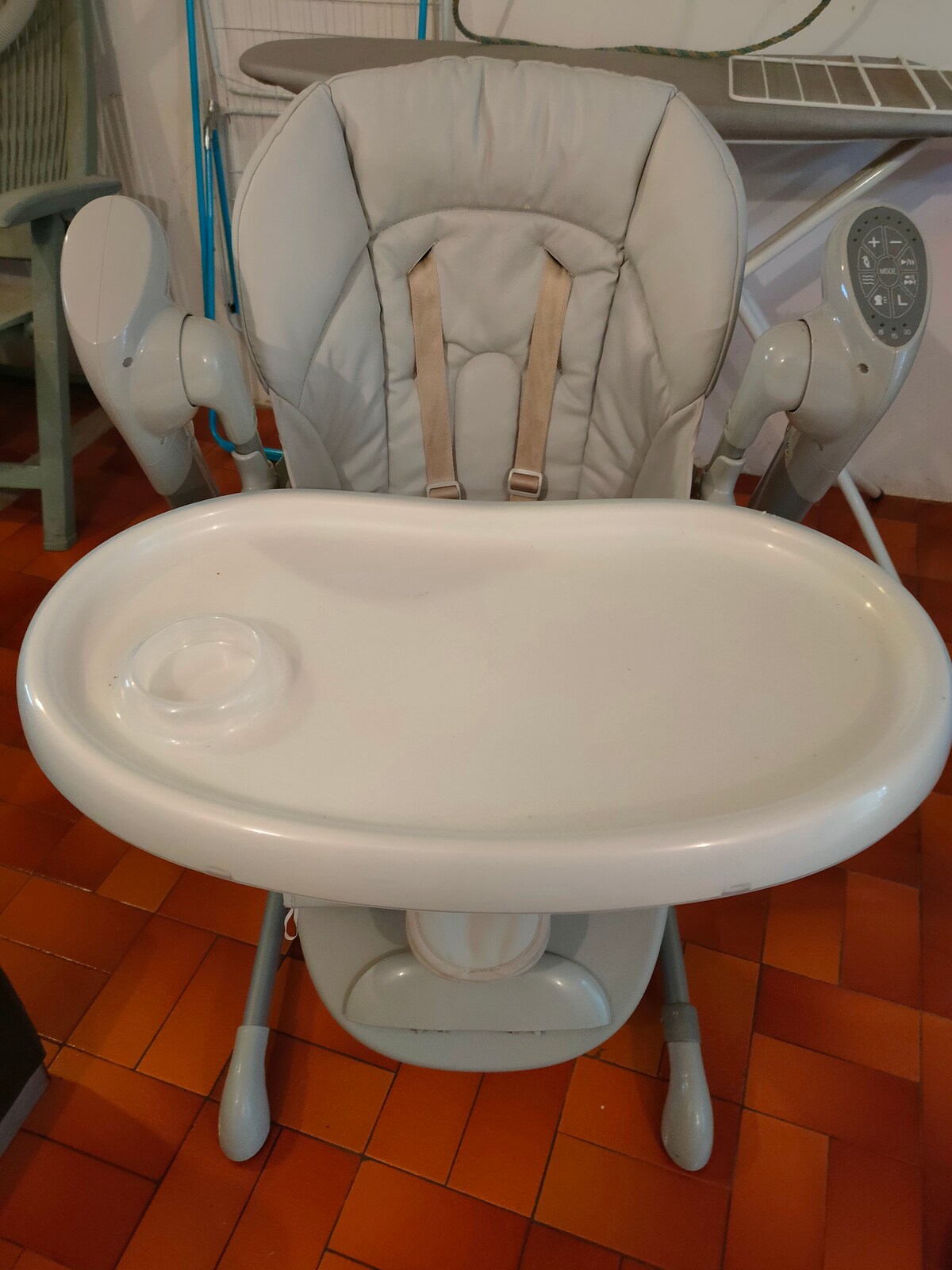A high chair is shown, featuring a soft, padded seat with safety straps. A large, detachable tray is positioned at the front, equipped with a small, circular indent. The chair's frame is sturdy, and additional elements are visible in the background.