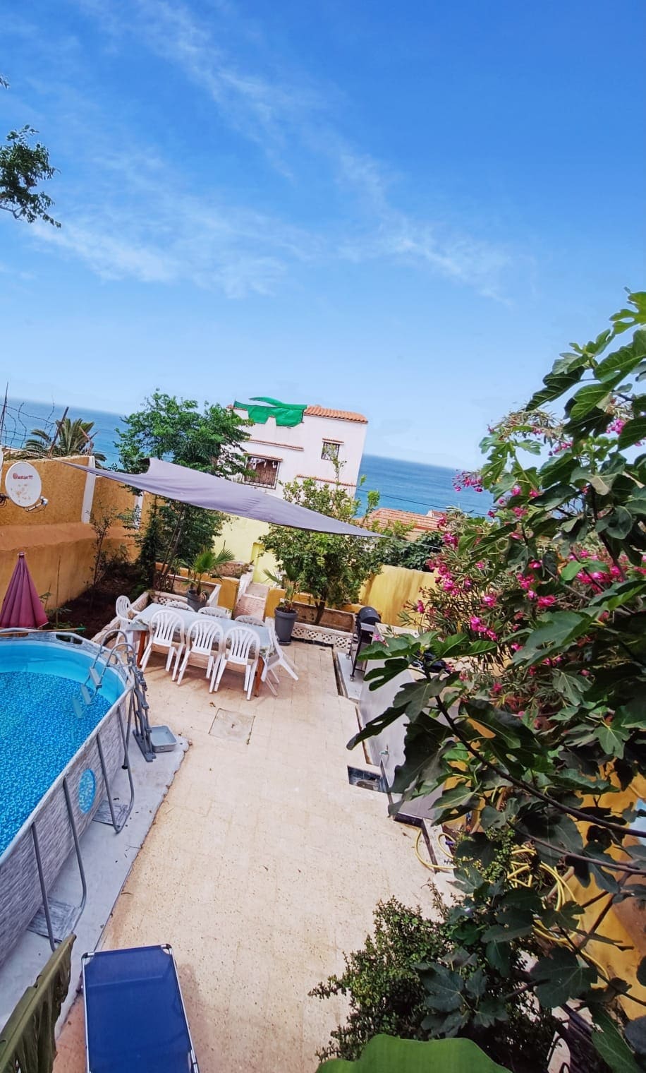 The outdoor area showcases a swimming pool surrounded by a tiled patio, with several chairs positioned nearby. Lush green plants and vibrant flowers frame the space, while a glimpse of the sea is visible in the background beneath a clear blue sky.