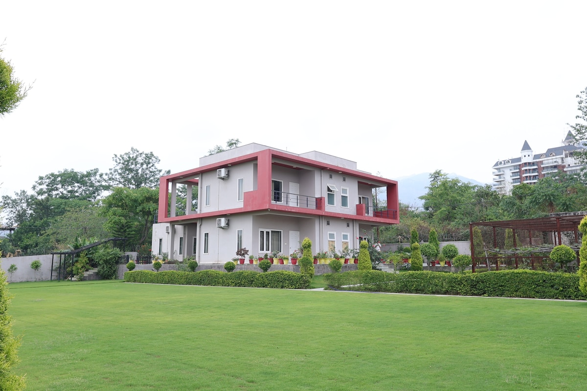 A modern two-story farmhouse is surrounded by a spacious lawn, featuring neatly trimmed greenery and vibrant flower beds. The building showcases a unique design with red accents and multiple air conditioning units, complemented by a serene backdrop of nature.