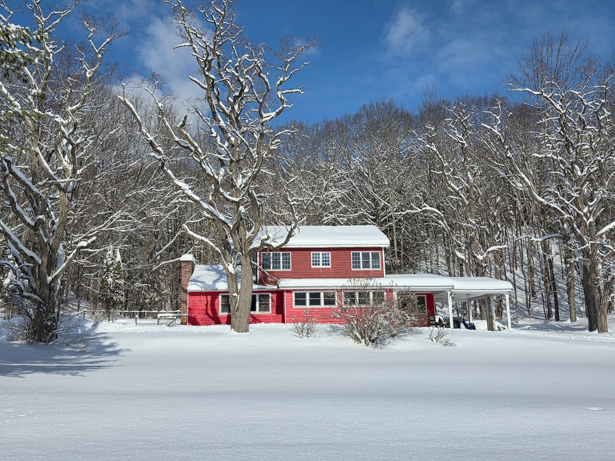 The vacation home is showcased in a winter landscape, surrounded by freshly fallen snow and tall trees. The two-story structure features a red exterior with white trim. A covered porch extends across the front, providing a cozy entry to the home, which is framed by a clear blue sky.