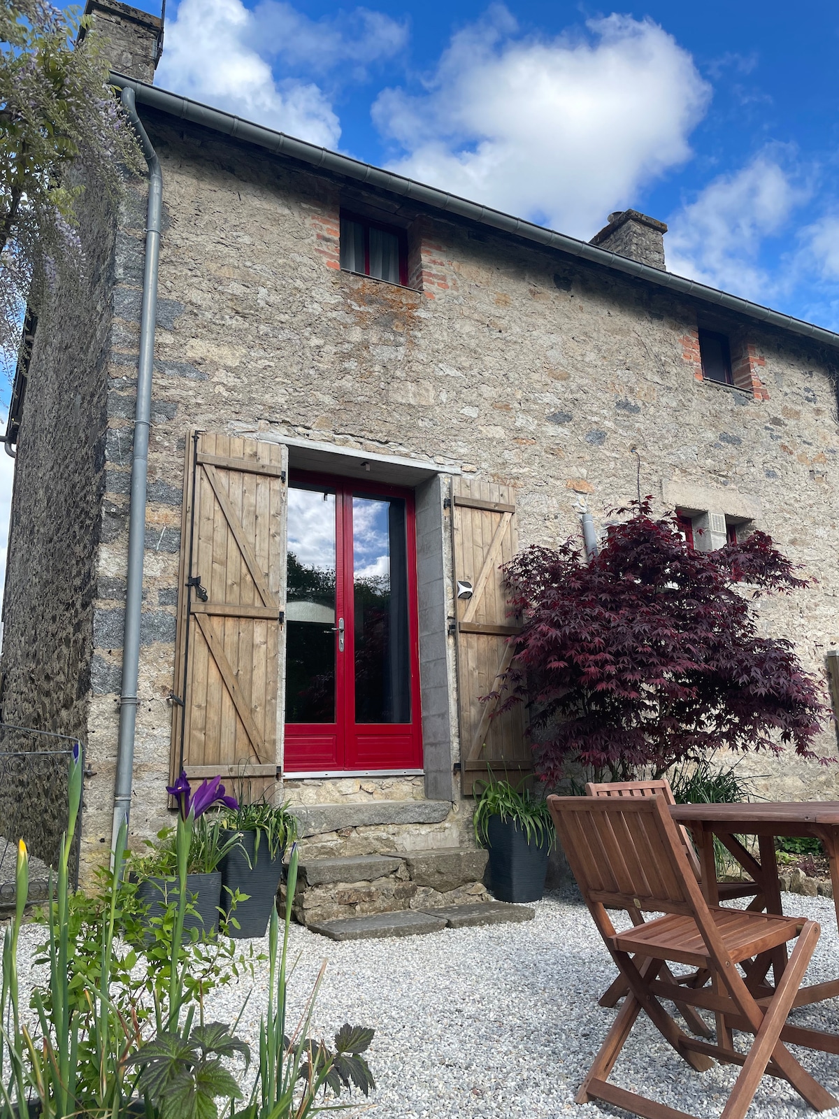 The exterior of a traditional stone house is depicted, featuring a striking red door framed by wooden shutters. Colorful plants are positioned beside a gravel pathway leading to the entrance, with a wooden dining table set nearby, creating a welcoming outdoor space.