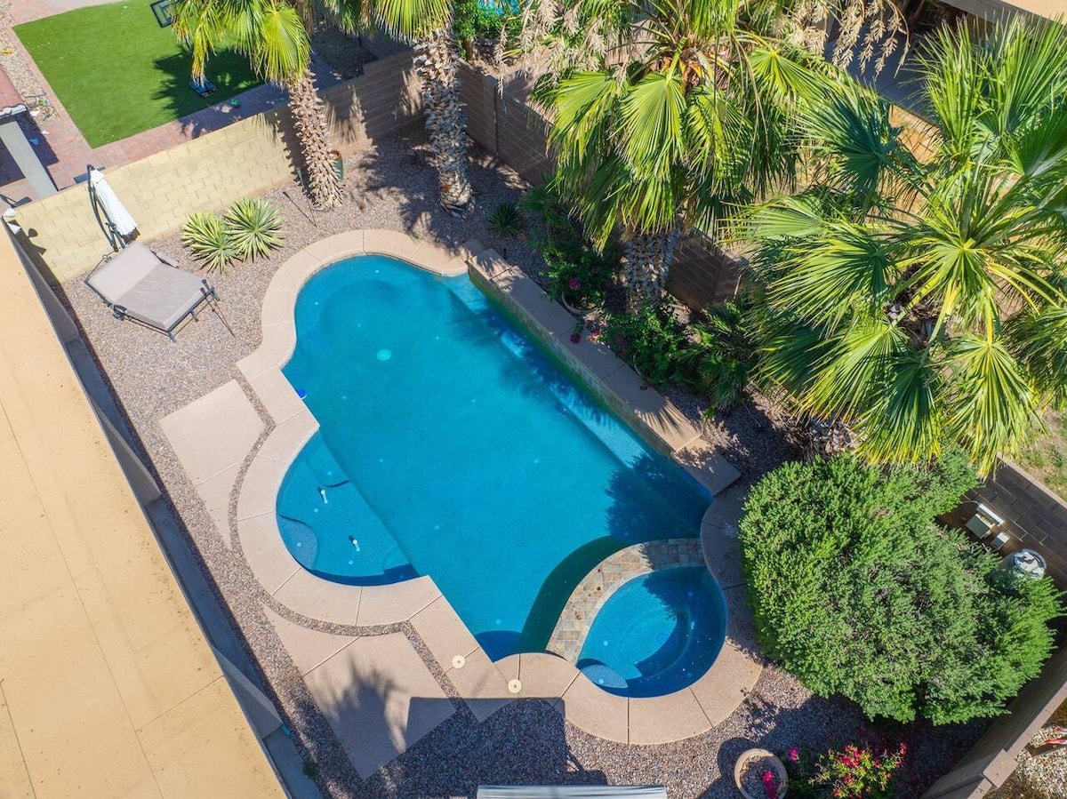 An aerial view of a private pool surrounded by landscaping, featuring palm trees and colorful shrubs. The pool is designed with a unique shape and includes a tanning shelf. The surrounding patio is paved and offers ample space for relaxation.