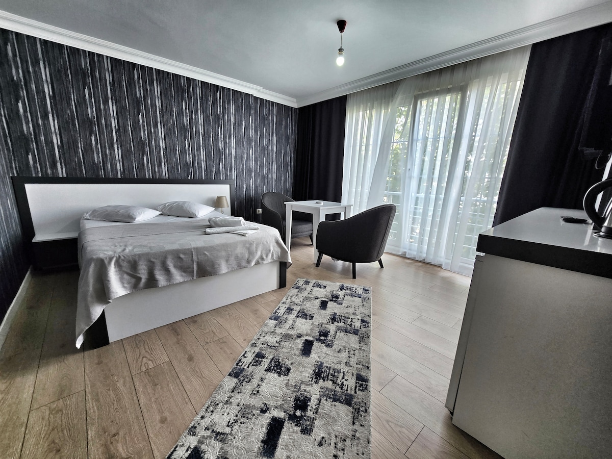 A modern bedroom is furnished with a spacious bed covered with white linens, and a light rug adds texture to the wooden floor. Natural light filters through sheer curtains, illuminating a small table and two chairs in the corner.