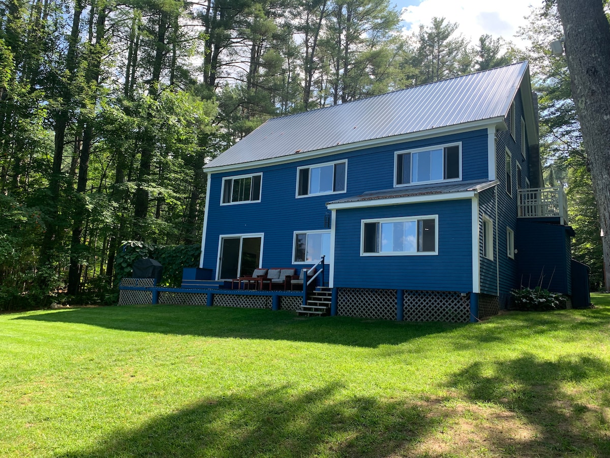 A two-story blue house is nestled among trees, featuring a large grassy area in the front. A deck with seating is visible, along with a staircase leading to the yard. Large windows allow for ample natural light, enhancing the home’s exterior appeal.