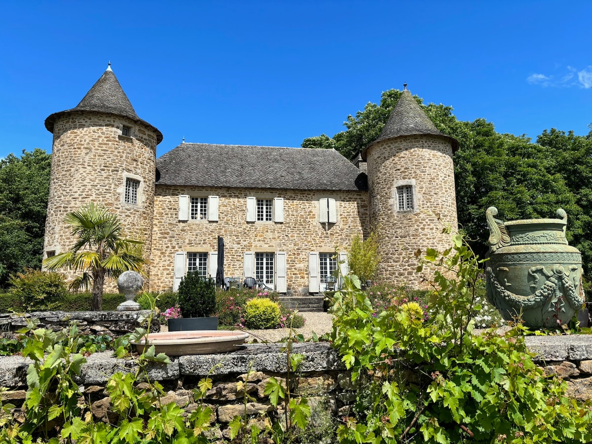 The stone façade of a castle displays round turrets, contrasting with a clear blue sky. Lush greenery surrounds the property, and a stone planter is visible in the foreground, adding to the garden's beauty. Sunlight illuminates the scene, enhancing the structure's charming architecture.
