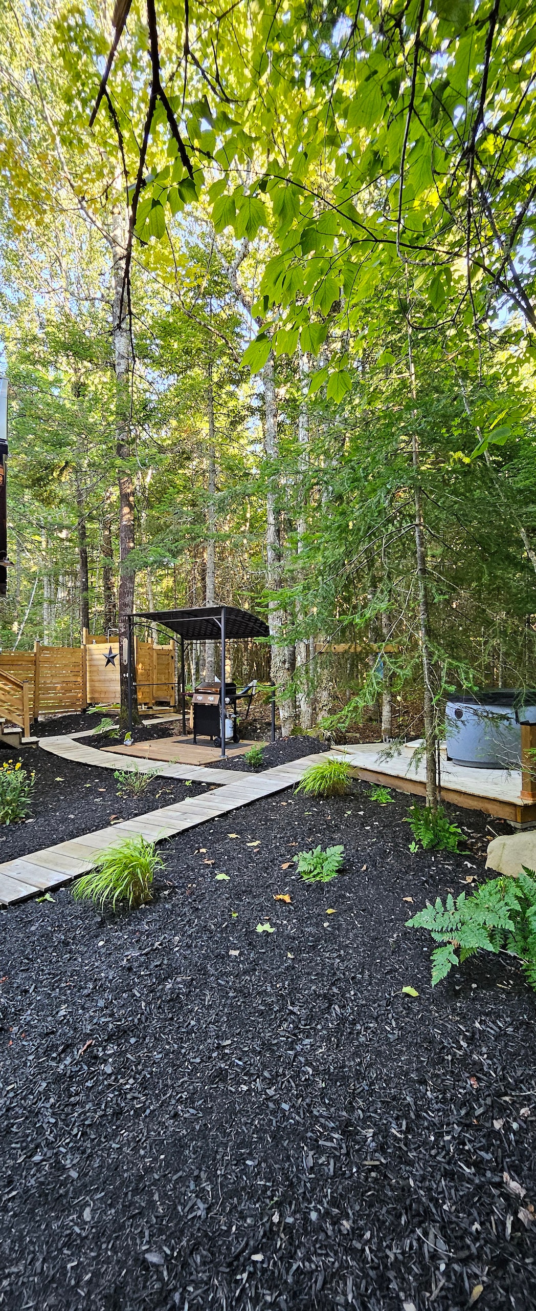 A shaded pathway leads through a natural landscape of lush greenery and black mulch, connecting various outdoor spaces. A gazebo is positioned along the path, with a hot tub nearby, surrounded by trees that provide a peaceful atmosphere.