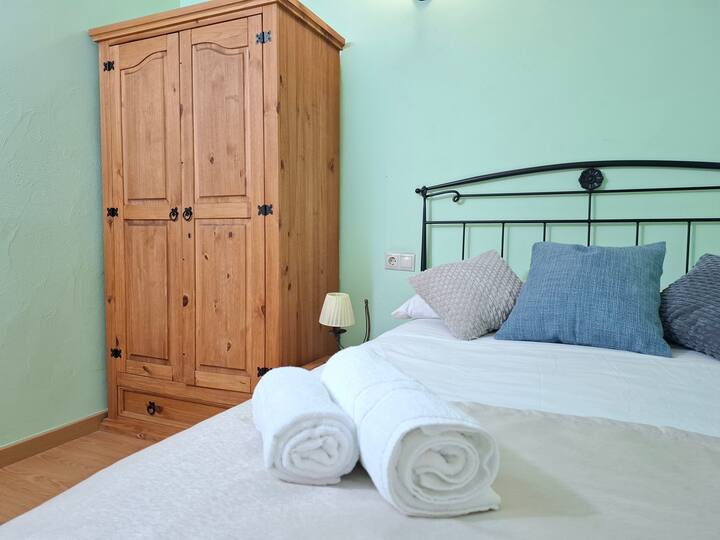ห้องพักพร้อมเตียงคู่มีตู้เสื้อผ้าไม้สไตล์ชนบทและหมอนตกแต่ง บรรยากาศสดใสด้วยโทนสีที่นุ่มนวลและอบอุ่นเหมาะสำหรับการพักผ่อนมีผ้าเช็ดตัวสีขาวพับอยู่เหนือเตียงอย่างสมบูรณ์แบบ