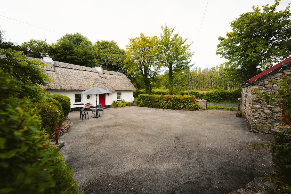 An outdoor view of the cottage features a gravel area surrounded by greenery, with trees and shrubs complementing the serene countryside. A table and chairs are positioned under an umbrella, providing a shaded spot to relax. The charming thatched roof of the cottage adds character to the scene.