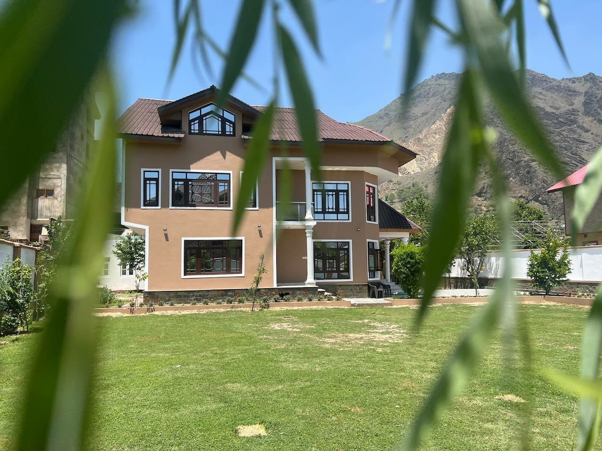 The exterior of the property is framed by green foliage, showcasing a spacious two-story building with multiple large windows. The facade features a blend of earthy tones, and a well-maintained lawn extends in front, with a backdrop of a mountainous landscape under a clear blue sky.