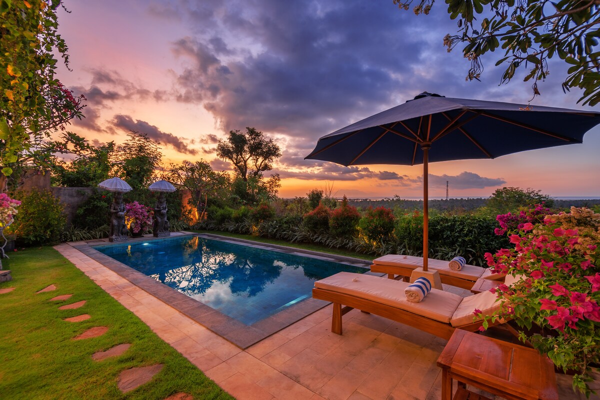 A private infinity swimming pool is surrounded by well-maintained gardens, featuring colorful flower arrangements. Two lounge chairs are positioned by the pool, beneath a large umbrella. A vibrant sunset casts warm hues across the sky, creating a serene setting for relaxation.