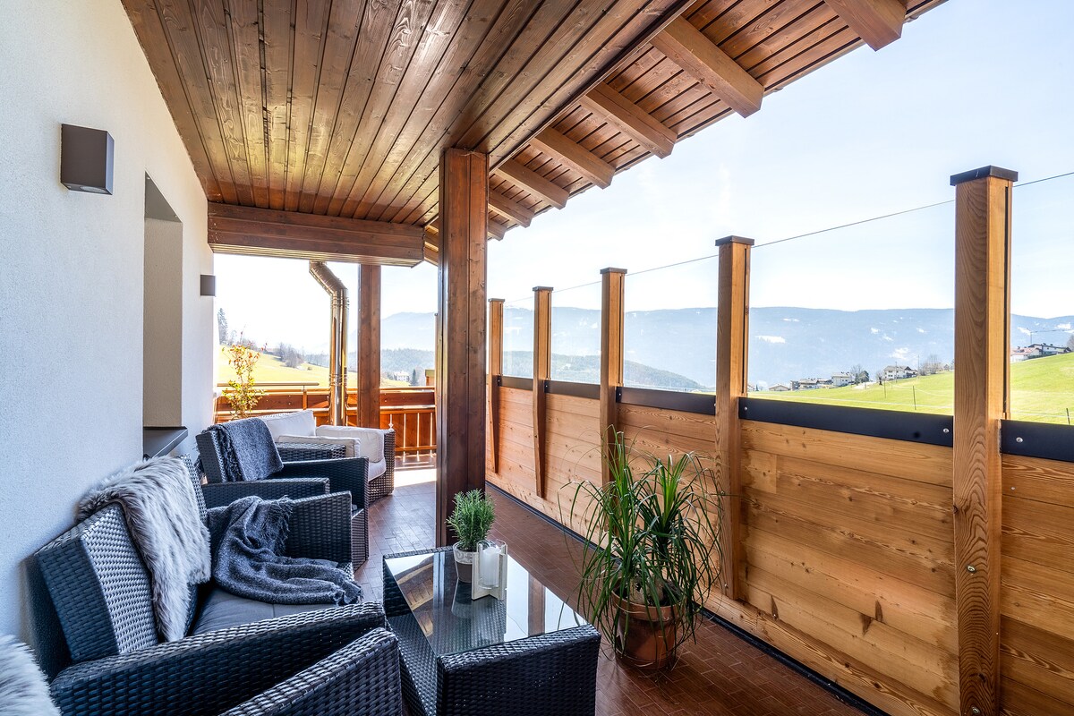 A spacious balcony features woven seating arrangements with soft cushions and a glass-topped coffee table. Surrounding railings are crafted from wood, framing expansive views of rolling mountains under clear skies. Potted plants add a touch of greenery to the serene outdoor space.