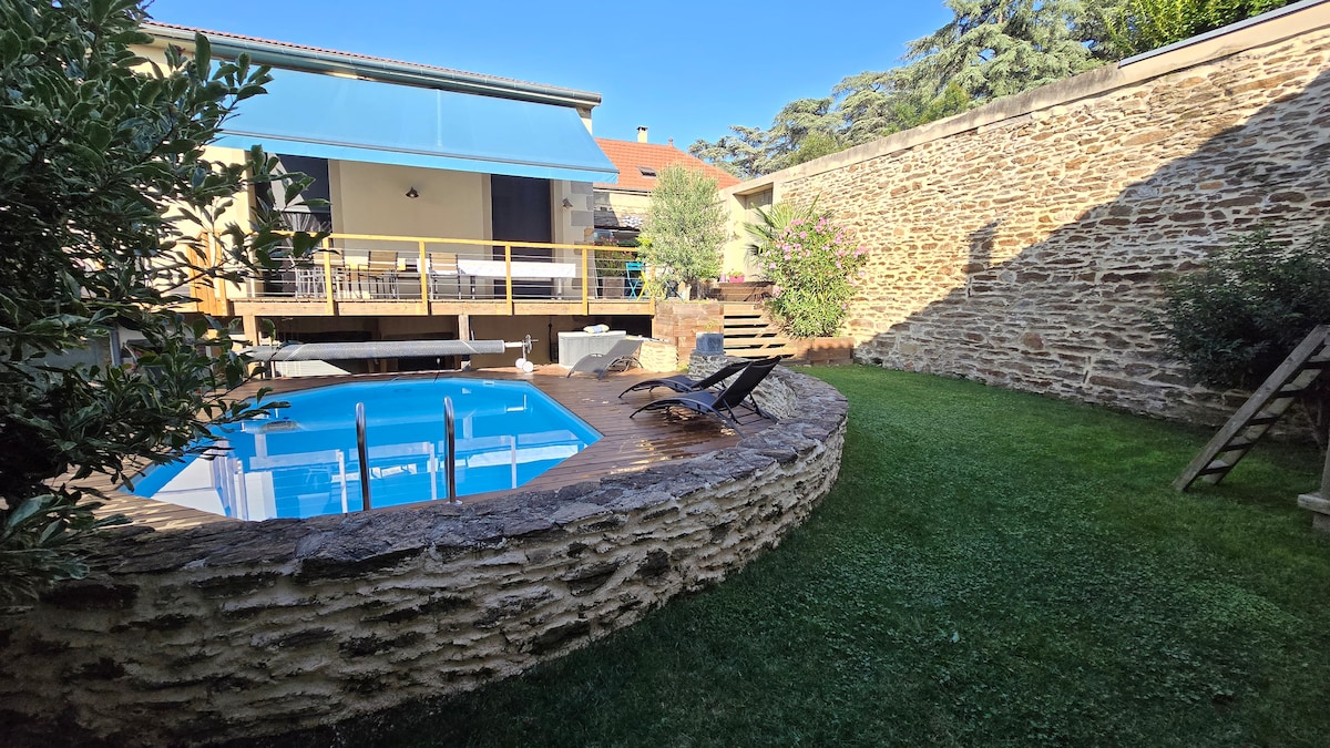An outdoor area features a private pool surrounded by a wooden deck and stone wall. A lawn is visible, complemented by a sliding door that opens to a terrace. Sun loungers are arranged nearby, providing a relaxing spot for guests.