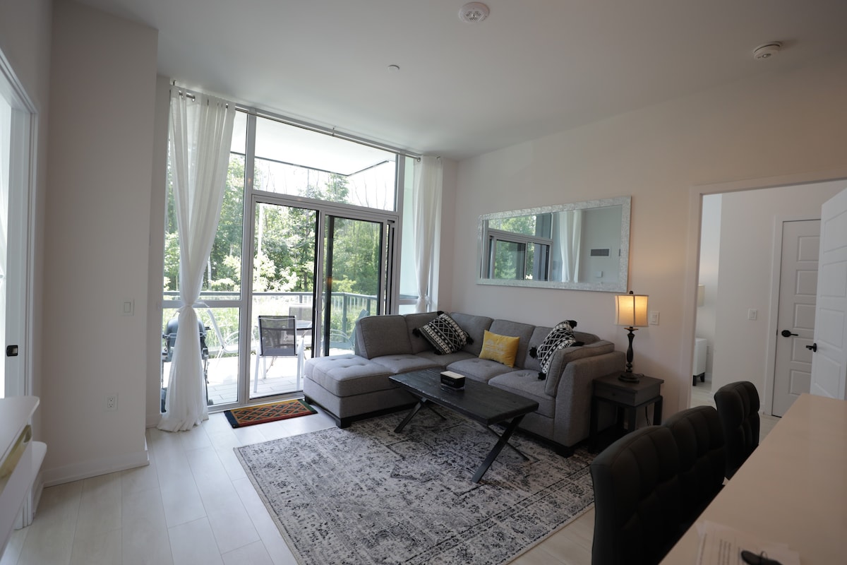 A well-lit living area is featured with large windows allowing natural light to enhance the space. A comfortable sectional sofa is positioned near a coffee table, with decorative pillows adding texture. A mirror reflects the outdoors, and a dining area is glimpsed in the background.