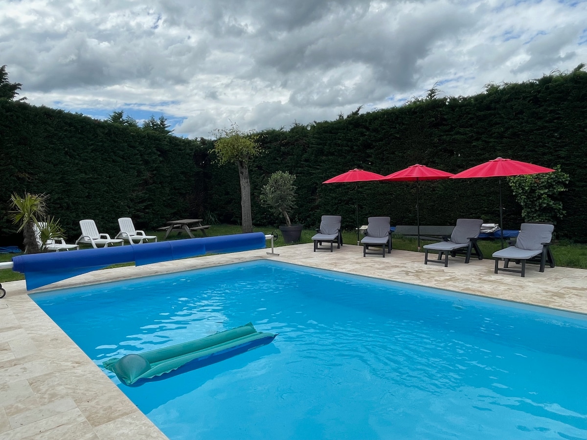 A well-maintained swimming pool occupies the center of the image, surrounded by sun loungers and vibrant red umbrellas. Lush greenery provides privacy along one side, while a seating area and picnic table can be seen in the background, creating a relaxing outdoor environment.