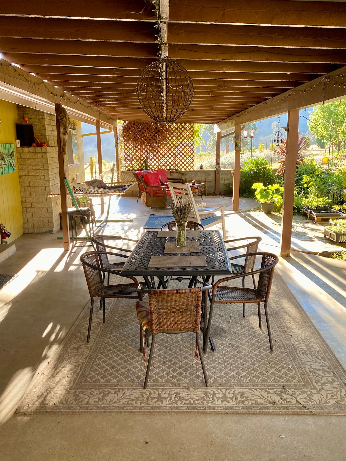 A shaded patio area features a large dining table surrounded by wicker chairs. Vibrant plants are arranged along the edges, and a cozy seating area with cushioned chairs is visible in the background, creating an inviting space for relaxation.