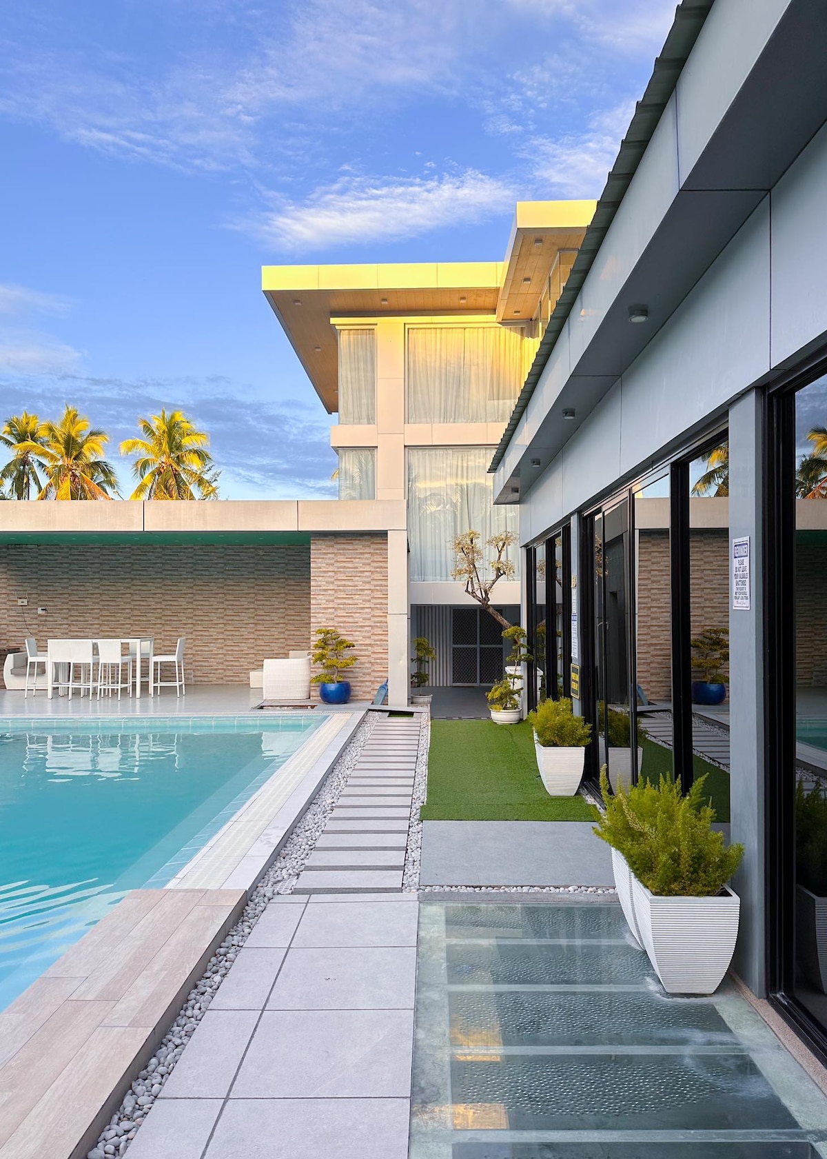 An outdoor area is shown featuring a clear swimming pool bordered by a tiled walkway. Lush potted plants are placed along the edge, and modern lounge furniture is arranged nearby. The building's expansive glass walls reflect the vibrant sky and surrounding palm trees.