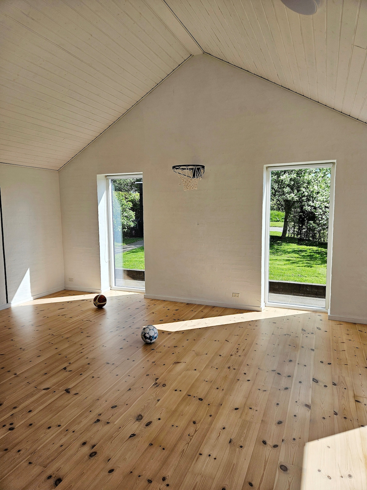 A spacious multi-purpose room is illuminated by natural light from large windows, showcasing a wooden floor. Two balls, one for basketball and one for soccer, are placed on the floor. The room offers an open view of the surrounding greenery.
