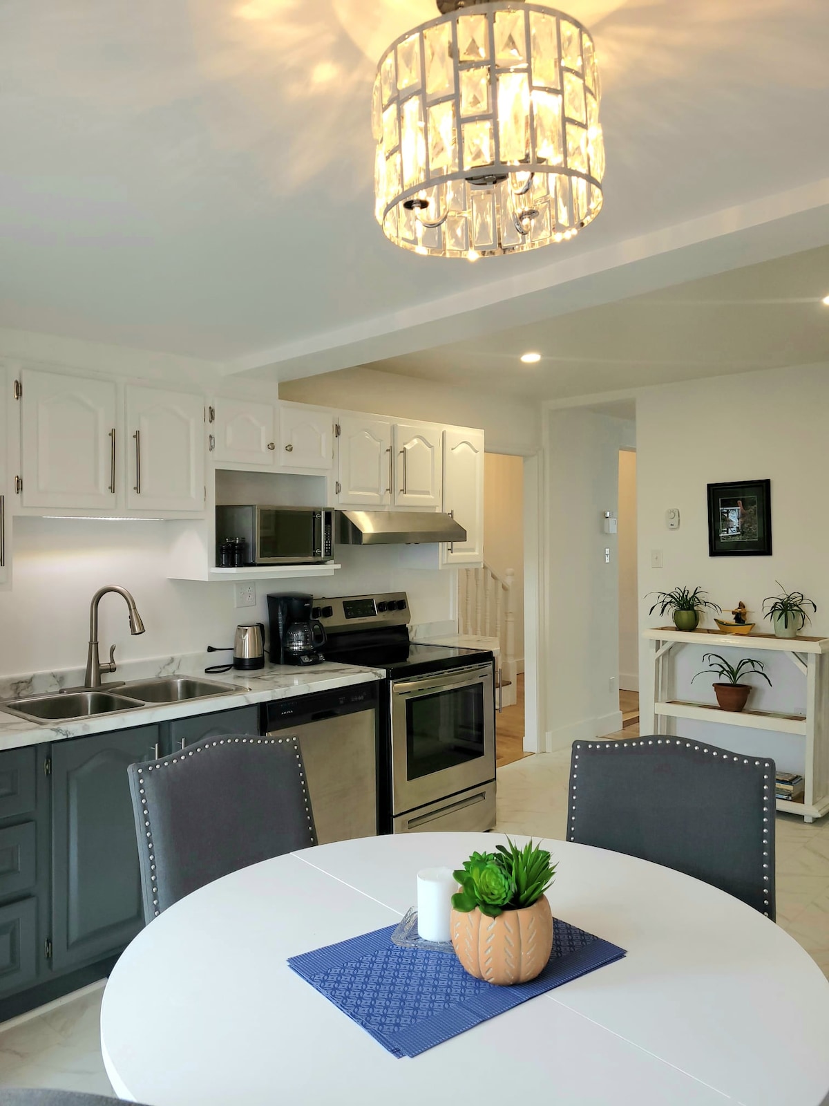 A modern kitchen space features white cabinetry and stainless steel appliances, including an oven and microwave. A round dining table, set with a decorative plant and light-blue placemat, invites casual meals. A stylish chandelier illuminates the room, enhancing its bright and airy feel.