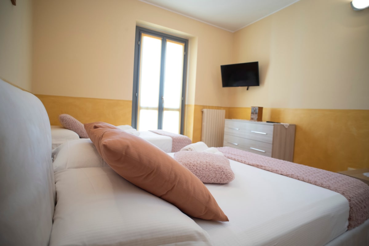 A bright bedroom features two single beds and one larger bed, each adorned with soft bedding and cushions. A television is mounted on the wall opposite a wooden dresser. Large windows allow natural light to fill the room, enhancing the warm color of the walls.