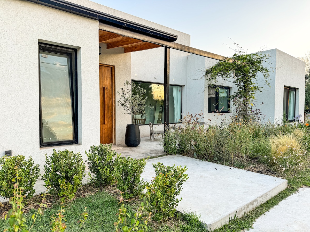 The entrance features a concrete pathway leading to a modern façade adorned with large windows and wooden accents. Surrounding greenery includes flowering plants and shrubs, creating a welcoming approach to the property.