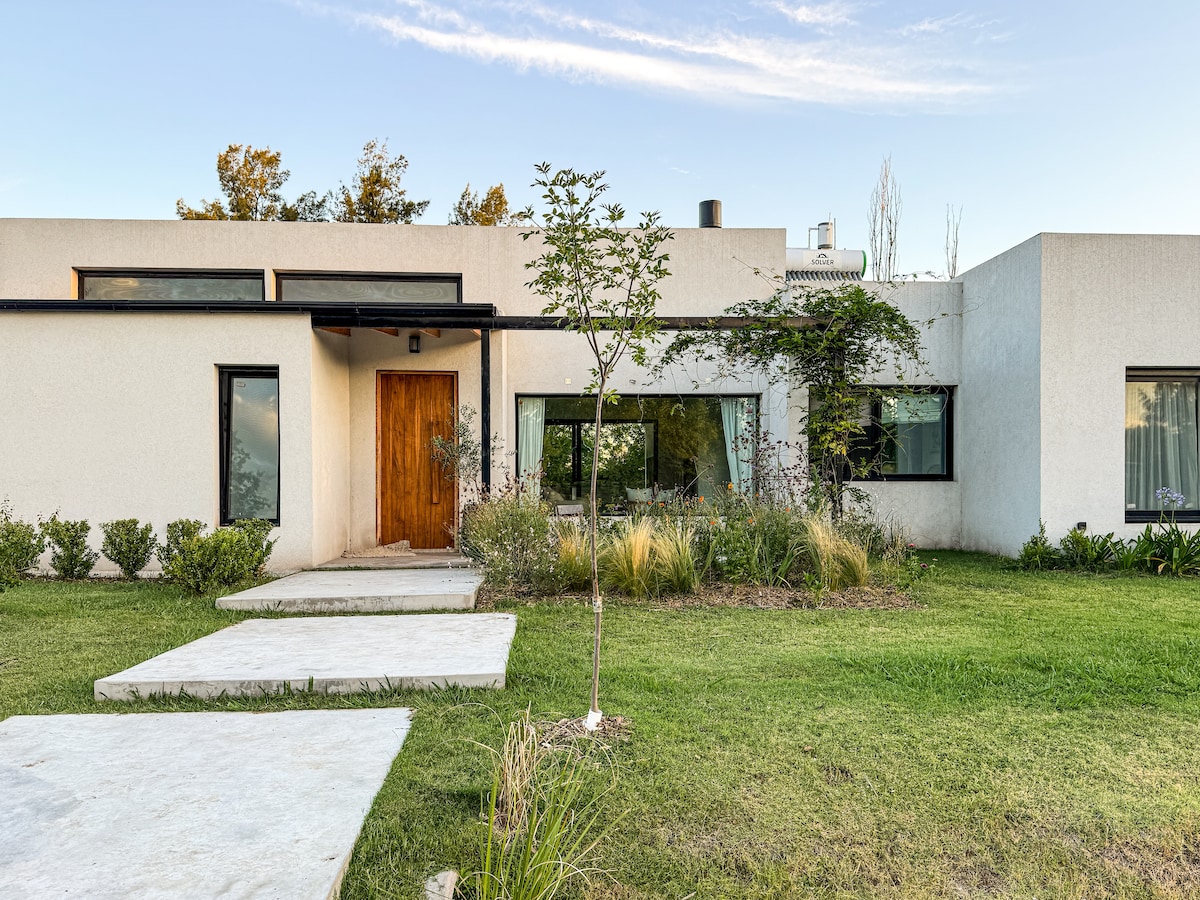 A modern single-story home is presented with a minimalist facade. A stone pathway leads to the welcoming wooden front door, surrounded by landscaped greenery and low-maintenance plants. Large windows allow natural light to fill the interior spaces, enhancing the connection with the outdoors.