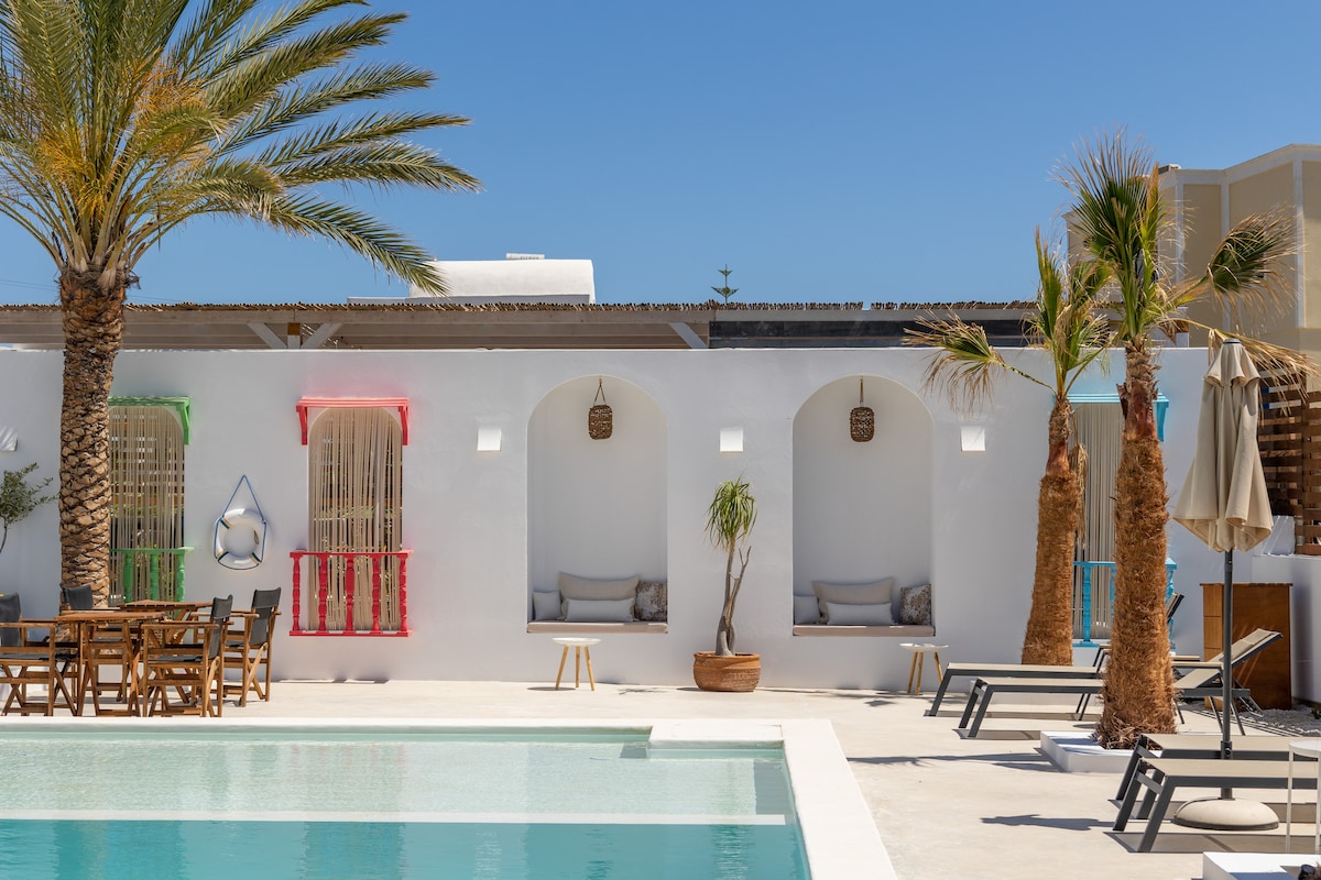 A modern exterior view of a Cycladic property features vibrant window accents in red and blue. Comfortable seating areas are positioned beneath the shaded overhangs. Lush palm trees and an inviting pool in the foreground enhance the outdoor space, while sun loungers are arranged around the pool area.