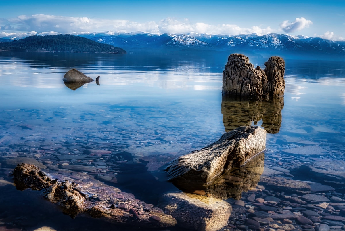 Calm waters reflect the surrounding mountainous landscape, with clear visibility of smooth stones beneath the surface. Driftwood and natural rock formations emerge from the lake, offering a serene view of the tranquil environment and distant snow-capped mountains under a bright blue sky.