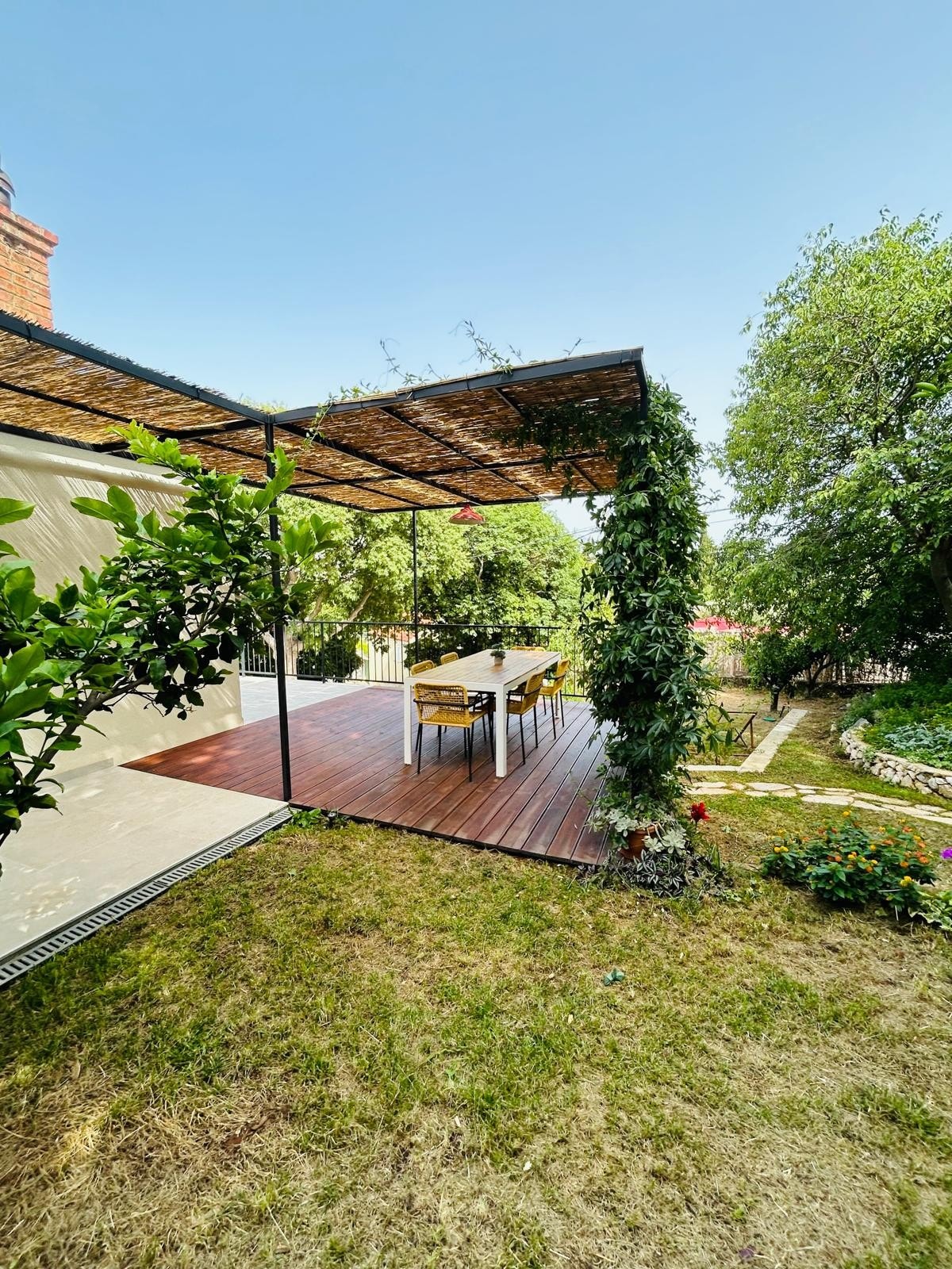 An outdoor dining area is sheltered by a sleek pergola, featuring a wooden table surrounded by several chairs. Lush greenery and flowering plants complement the scene, with a well-maintained yard providing a natural backdrop for relaxation and meals in a tranquil atmosphere.