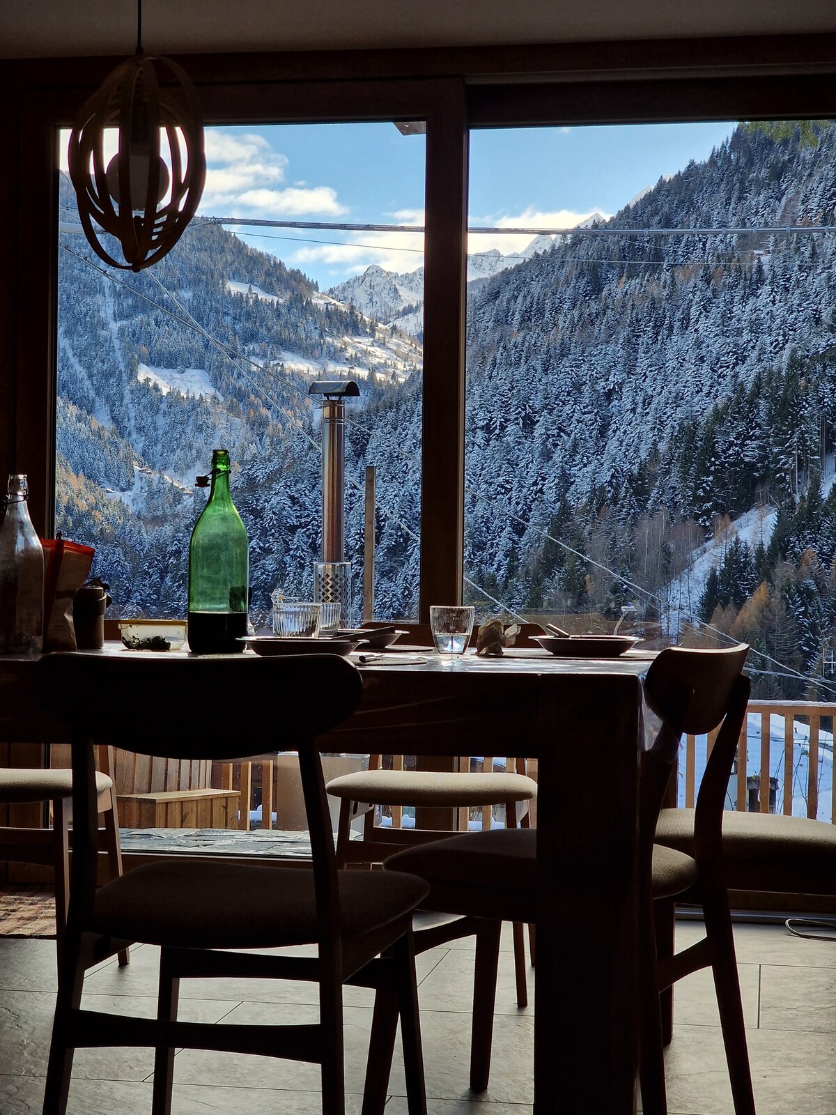 A warm dining area features a wooden table set for a meal, with glassware and a green bottle present. Large windows reveal snow-covered mountains and a clear blue sky, creating a serene backdrop that enhances the inviting atmosphere of the space.