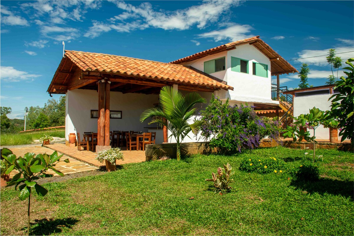 A charming two-story cabin is showcased, featuring a terracotta tiled roof and wooden beams. The surrounding area is filled with green grass and various plants, providing a natural setting. A dining area with wooden tables is visible on the patio, offering outdoor seating.