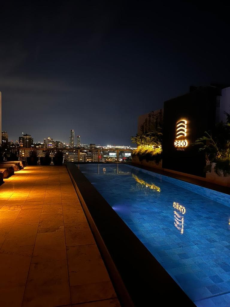 An illuminated rooftop infinity pool reflects the night sky, surrounded by a serene ambiance. The city skyline is visible in the background, with lights twinkling from various buildings. Soft lighting along the pool's edge provides a calm atmosphere for relaxation.
