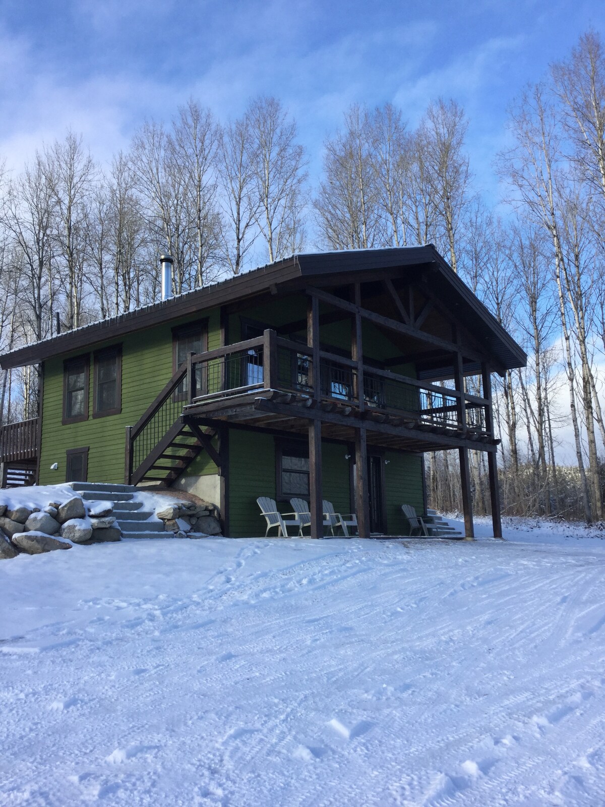 A green chalet stands amidst snow-covered surroundings, featuring a spacious deck with steps leading to the entrance. Four chairs are positioned on the deck, and tall trees are visible in the background under a partly cloudy blue sky.