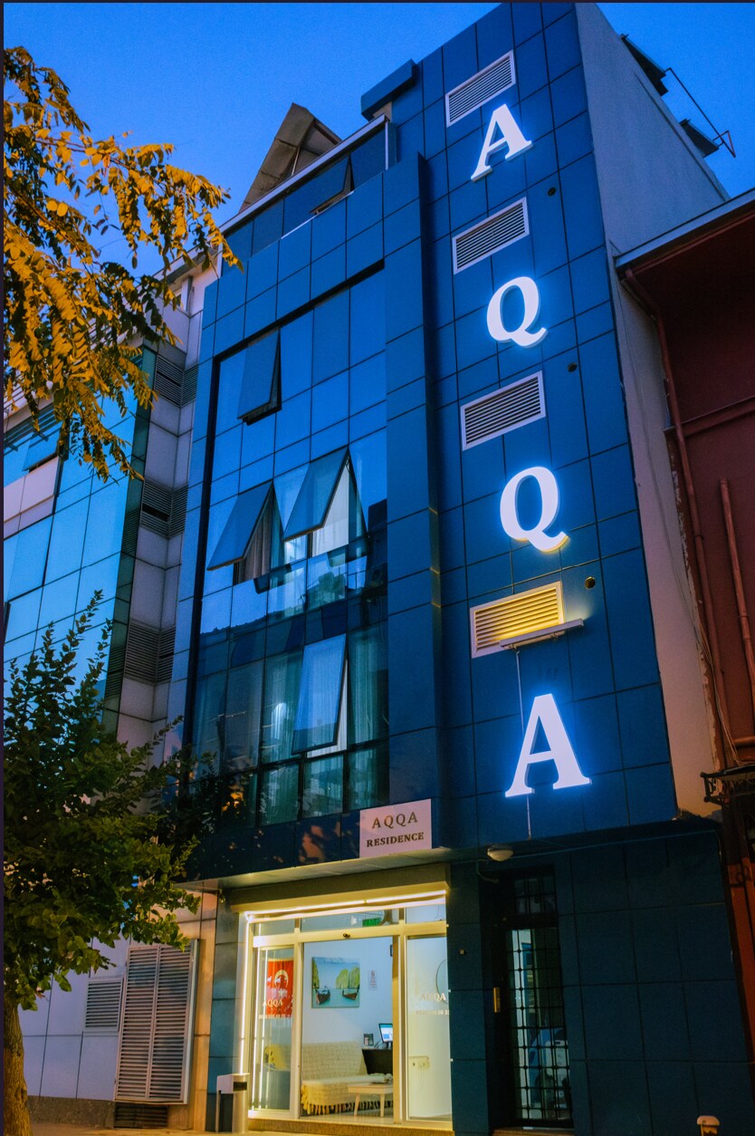 The exterior of the building is seen at dusk, featuring a modern design with large glass windows. The illuminated sign displaying 'AQQA' is prominently positioned on the front, enhancing visibility. Surrounding greenery adds a touch of nature to the urban setting.