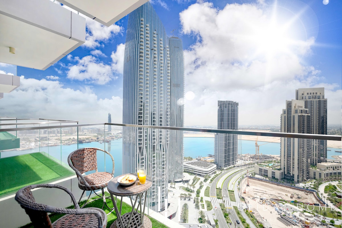 A balcony overlooks a stunning urban landscape, showcasing tall skyscrapers and the shimmering water of the creek below. Two wicker chairs and a small table are set for two, adorned with a glass of orange juice and a plate. Bright sunlight accentuates the scene.
