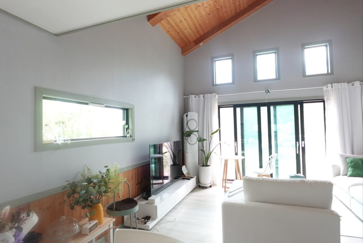 A bright living area features large glass doors opening to the outdoor space, allowing natural light to fill the room. A sofa and TV are positioned for relaxation, while a plant adds a touch of greenery. The wooden ceiling provides an open and airy feel.