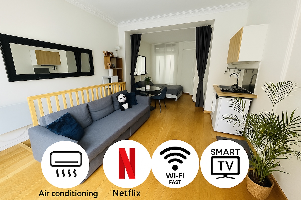 A spacious living area features a comfortable dark gray sofa against a white wall. A Samsung Smart TV is mounted above a kitchen area with modern appliances. A small bed is visible in the background, enhancing the functional layout. Light wood flooring complements the bright, airy atmosphere.