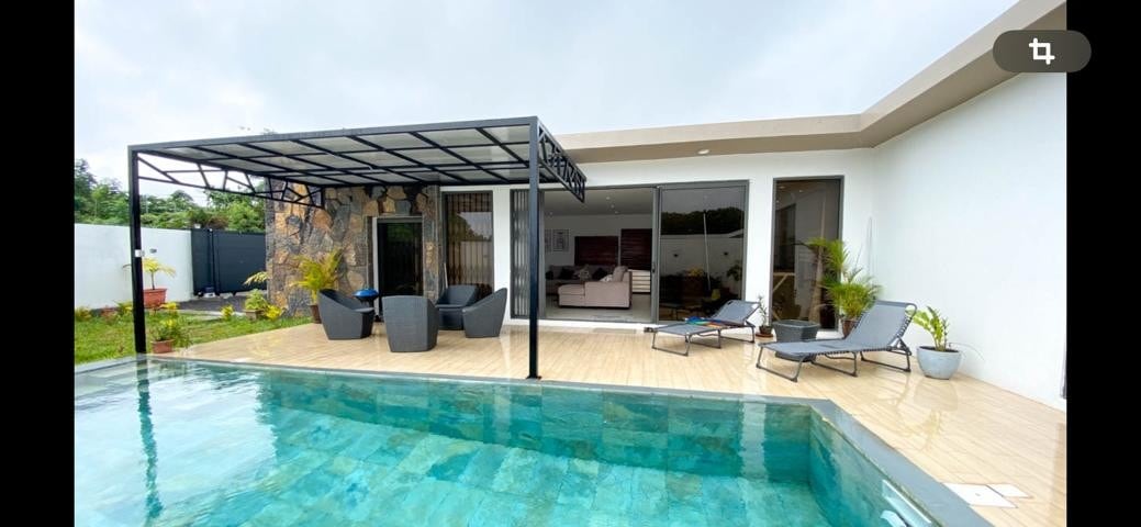 A private swimming pool reflects natural light, bordered by a wooden deck. Modern outdoor seating is arranged under a shaded pergola, with lounge chairs positioned nearby. The villa's expansive glass doors provide a view of the inviting living space, merging indoor and outdoor living.