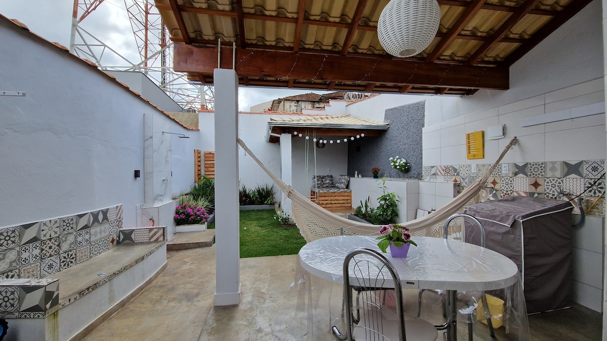 An outdoor area features a circular dining table surrounded by transparent chairs. A hammock is suspended in a shaded corner, while vibrant plants and flowers add greenery to the space. A decorative wall with patterned tiles enhances the inviting atmosphere.