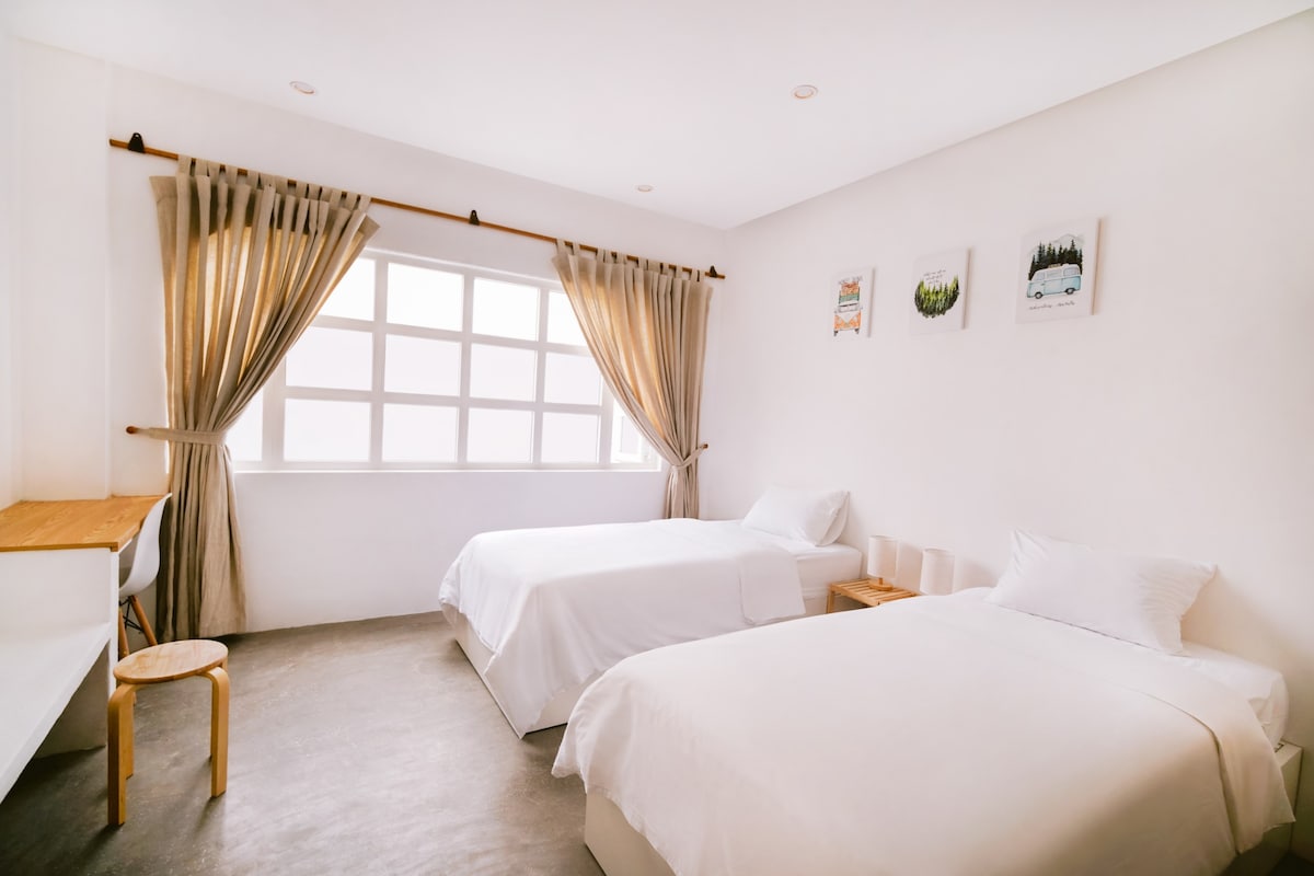 A softly lit room features two spacious beds dressed in white linens. Natural light fills the space through a large window framed by sheer curtains. Simple wall art adds a touch of color, while a small desk and chair provide a functional work area.