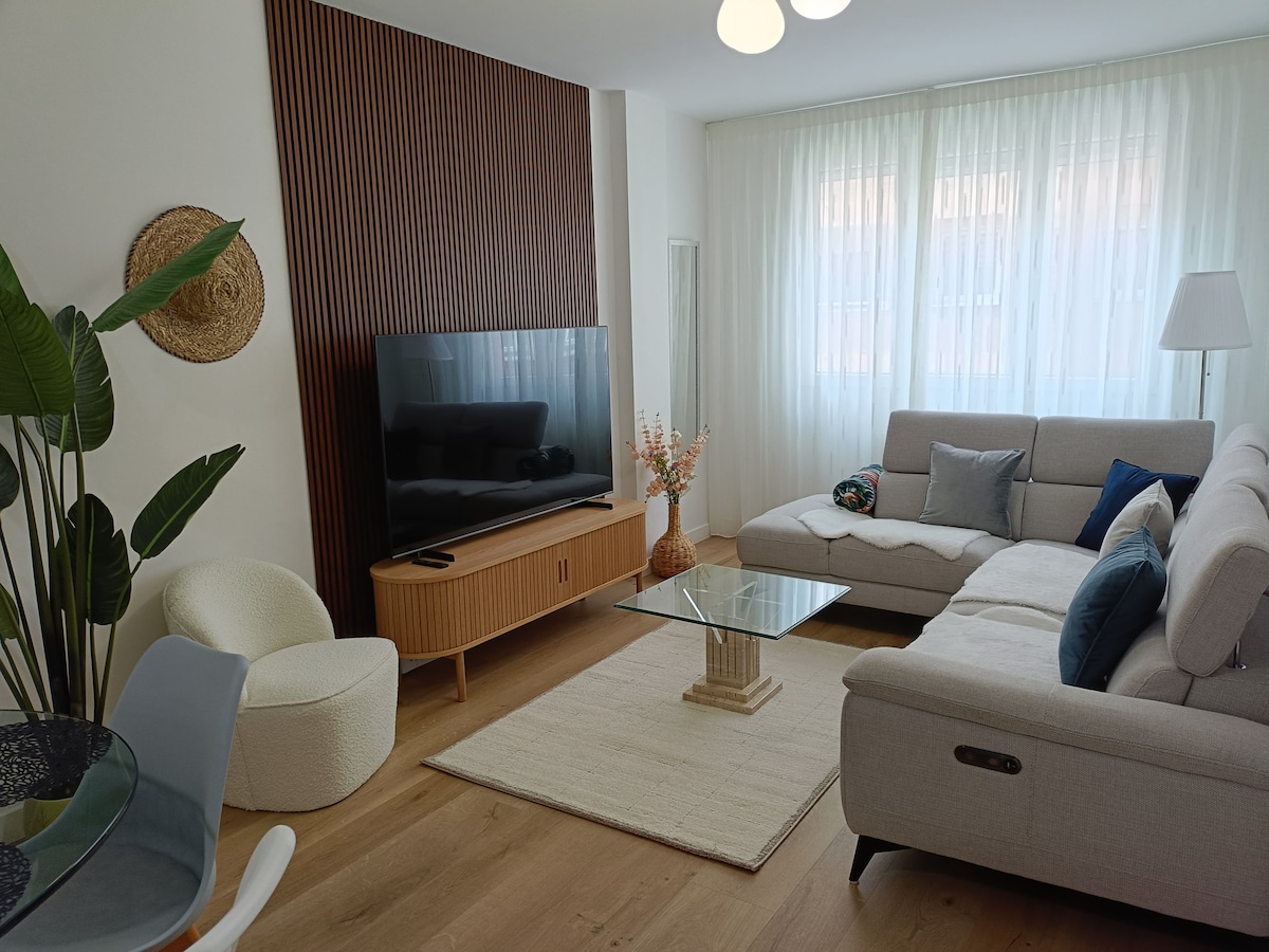 A spacious living area is presented with a light-toned sectional sofa and a glass coffee table on a soft area rug. Natural light filters through sheer curtains, highlighting a modern entertainment unit with a large television. Decorative plants and a tasteful arrangement of flowers add subtle accents.