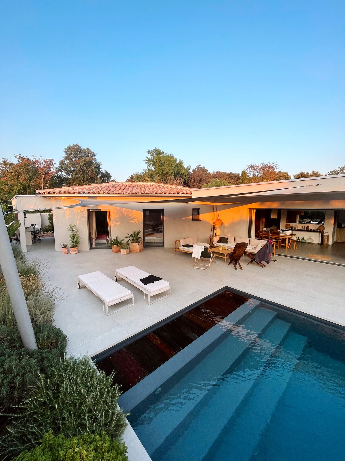 A spacious outdoor area features a rectangular pool partially shaded by a terrace. Comfortable loungers and seating arrangements are positioned thoughtfully around the pool. The sunset light casts a warm glow over the detailed landscape and the nearby home with its modern architecture.