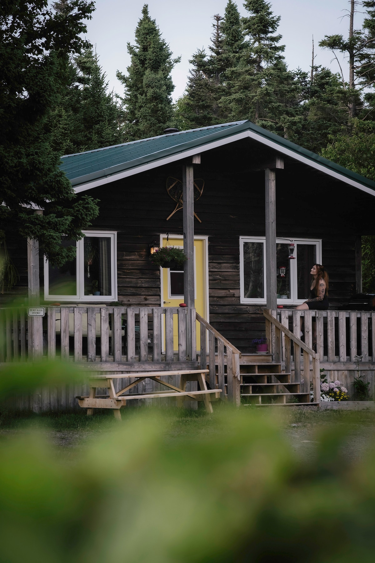 A rustic cabin sits among lush greenery, featuring a welcoming yellow door and a covered front deck. Sturdy steps lead up to the entrance, and a picnic table is positioned nearby, inviting relaxation in the natural surroundings.